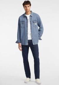 Camisa de denim azul claro con dos bolsillos delanteros, cierre de botones y mangas largas. Combinada con unos vaqueros azul oscuro y zapatillas blancas.