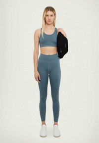 Brassière de sport turquoise et leggings assortis en tissu extensible, associés à des baskets blanches. Une veste noire est posée sur l'épaule.