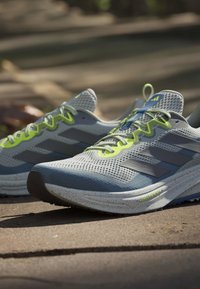 Chaussures de course gris clair et bleu avec des accents jaune néon et en mesh, placées sur un trottoir en extérieur.