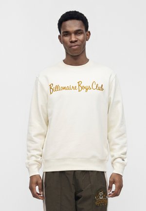 Jeune homme portant un sweat-shirt crème avec le texte « Billionaire Boys Club » en doré et un pantalon foncé avec un logo assorti sur la poche.