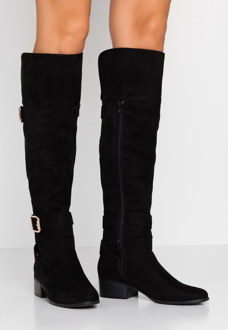 Botas negras de ante por encima de la rodilla con cremallera lateral, dos correas con herrajes rectangulares y tacón cuadrado. Textura suave.