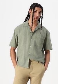 Camicia a maniche corte, con bottoni, in tessuto verde pallido con una texture, dotata di tasca sul petto e vestibilità rilassata. Abbinata a pantaloni beige.