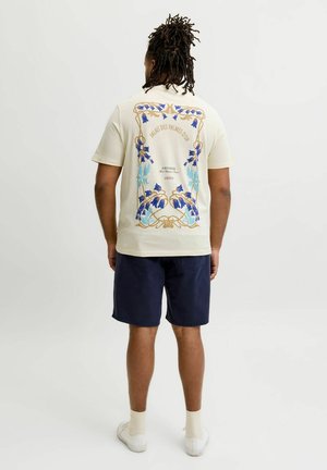 Bărbat cu dreadlocks purtând un tricou crem cu un desen floral albastru și text, pantaloni scurți bleumarin, șosete albe și adidași albi, stând cu fața spre spate.