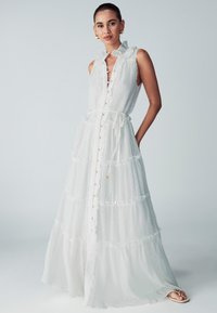 Abito maxi bianco a strati con bottone frontale e scollatura arricciata. Realizzato in tessuto leggero, con dettagli dorati e una cintura in vita.