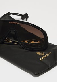 Occhiali da sole con montatura nera oversized, lenti marroni sfumate, dettagli dorati e una custodia nera texturizzata con logo.