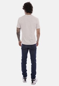 Uomo con capelli ricci che indossa una maglietta bianca semplice, jeans blu scuro, sneakers bianche e tatuaggi sul braccio sinistro, in piedi di spalle.