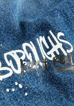 Tessuto denim di un blu profondo con una finitura testurizzata. Presenta un testo bianco prominente in stile graffiti e un testo in nero con una stampa grafica.