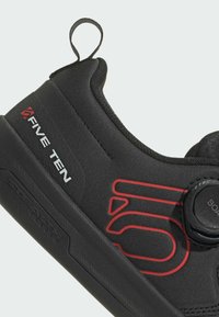 Musta kiipeilykengä, jossa on punaisia yksityiskohtia ja säädettävä suljin. Valmistettu synteettisestä materiaalista, ja siinä on kuvioitu pohja pidon parantamiseksi. Varustettu brändin logolla.