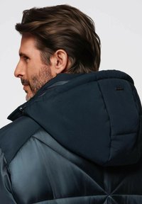 Marinblå puffjacka med en texturerad huva och kviltade detaljer, med både släta och matta ytor. Svart logotag synlig på baksidan.