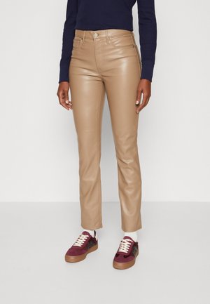 Personne portant un pantalon en similicuir beige, un haut à manches longues sombre, des chaussettes blanches et des baskets bordeaux avec des lacets blancs, debout devant un fond uni.