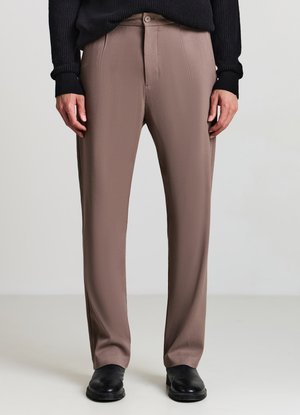 Homme portant un pantalon marron sur mesure, un pull noir et des chaussures en cuir noires, debout devant un fond clair uni.