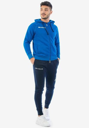 Giovane uomo in piedi con le mani nelle tasche, indossa una felpa con cappuccio blu con zip, pantaloni sportivi blu scuro con loghi e sneakers bianche su sfondo bianco.
