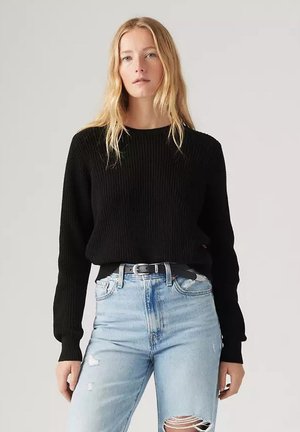 Suéter negro de punto con textura acanalada, cuello redondo y mangas largas, combinado con jeans claros desgastados y un cinturón negro.