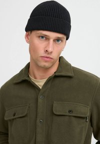 Chaqueta de forro polar verde con botones en la parte delantera, bolsillos de parche y gorro de canalé negro. El modelo tiene ojos azules y una camiseta neutra debajo.