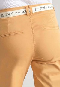 Pantaloni arancioni chiari con una texture liscia e una vita elastica con il testo "LE TEMPS DES CERISES". Include due tasche sul retro.