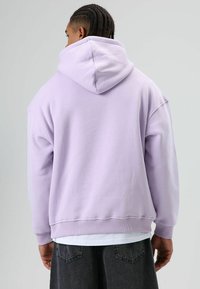 Lavendel oversized hoodie met een capuchon met trekkoord, geribbelde manchetten en zoom, gemaakt van zachte stof, gedragen over een wit T-shirt en grijze broek.
