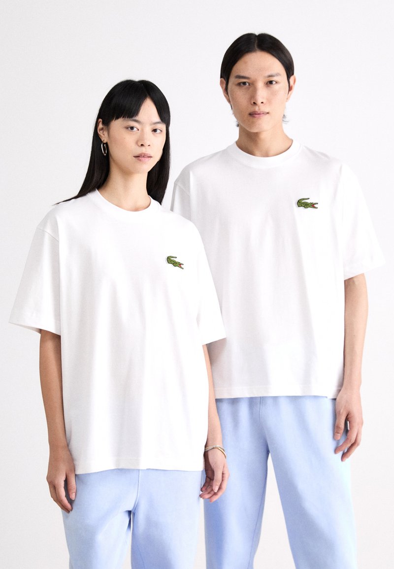 Camisetas de algodón blancas con escote redondo, que presentan logotipos de cocodrilo bordados en verde en el lado izquierdo. Combinadas con pantalones azul claro.