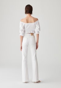 Haut blanc court à épaules dénudées avec manches bouffantes, associé à un jean blanc évasé taille haute avec une finition texturée et des coutures discrètes.