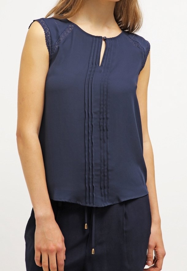 Blouse sans manches bleu marine avec finition en dentelle aux épaules, plis verticaux à l'avant, encolure goutte avec fermeture bouton, portée avec un pantalon bleu marine assorti.