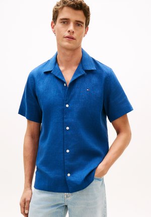 Junger Mann trägt ein kurzärmeliges blaues Hemd mit Knöpfen und helle blaue Jeans, steht mit einer Hand in der Tasche vor einem einfarbigen Hintergrund.