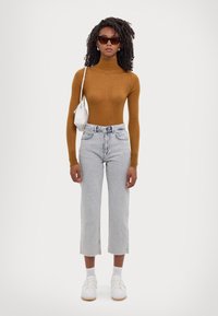 Top de cuello alto marrón, vaqueros cortos de mezclilla azul claro con dobladillo deshilachado, zapatillas blancas y bolso pequeño blanco. Los accesorios incluyen gafas de sol y pendientes.