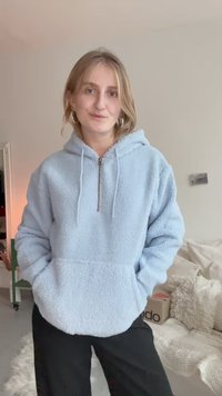Hoodie en peluche bleu clair avec un col zippé, des poches avant et une coupe décontractée. Porté avec un pantalon noir sur un fond neutre.