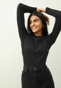 Mujer sonriendo con los brazos levantados detrás de la cabeza, vestida con una camisa negra de manga larga y jeans oscuros de tiro alto sobre un fondo liso.