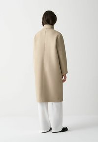 Beige lång kappa med hög krage, tillverkad av mjukt material, med rak design och en enda knappdetalj vid nederkanten. Bärs med vita byxor.