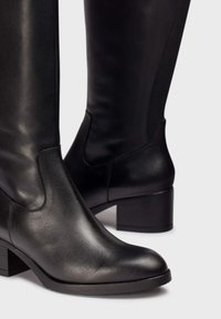 Botas de cuero negro hasta la rodilla con un acabado texturizado, costuras laterales curvadas y un tacón bajo cuadrado. Superficie lisa y diseño elegante.