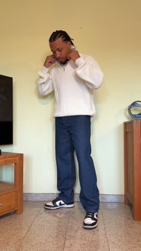 White knitted sweater with a high collar, paired with dark blue jeans and black and white sneakers. The setting features light-colored walls.