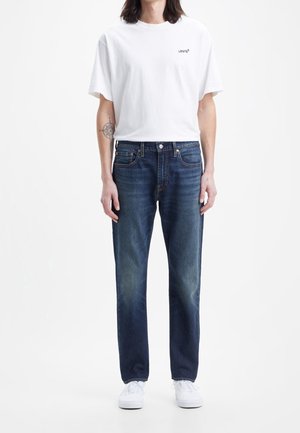 T-shirt bianco in cotone con un piccolo logo nero. Jeans blu scuro a gamba dritta con leggera scoloritura, abbinati a sneakers bianche.