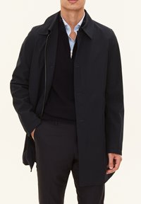 Marinblå lång kappa med knapp- och dragkedjeslutning, prydd med krage och sidofickor, över en svart tröja och en ljusblå skjorta.