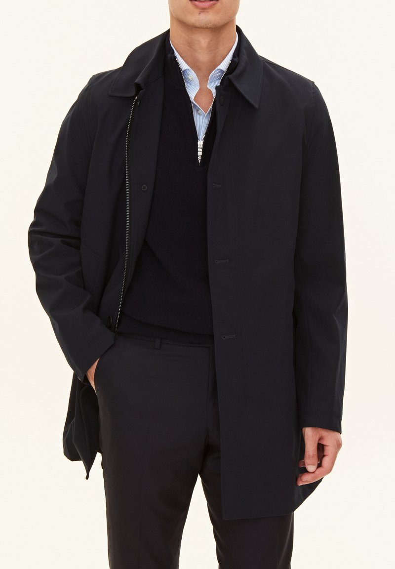 Marinblå lång kappa med knapp- och dragkedjeslutning, prydd med krage och sidofickor, över en svart tröja och en ljusblå skjorta.