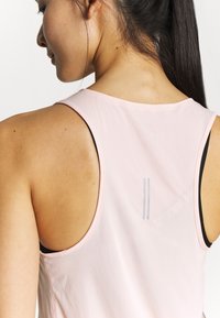 Mujer joven con cabello largo y oscuro que lleva una camiseta de deporte sin mangas de color rosa claro con franjas reflectantes en la parte superior de la espalda.