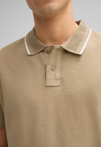Beige Poloshirt mit strukturiertem Kragen und weißen Akzenten, zwei Knopf-Leiste und einer weichen Stoffoberfläche.
