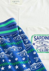 Witte T-shirt met een zak en de tekst "SARDINE CLUB" in grafische vorm. Blauwe shorts met een wit visprint en een groen golvend patroon, afgewerkt met een gestreepte tailleband.