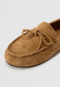 Brown suede moccasin shoe with rounded toe, leather laces, and subtle stitching detail. Features a flat rubber sole for comfort.
