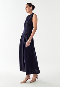 Mornarice modra brezrokavna obleka z zategnjeno pasom, nabranim krilom in subtilnimi gumbi. V kombinaciji s bež okrašeno obuvalo.