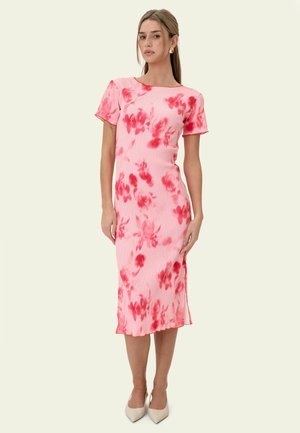 Kvinde står iført en lyserød midi-kjole med røde blomsterprint, korte ærmer og beige spidse hæle mod en ensfarvet baggrund.