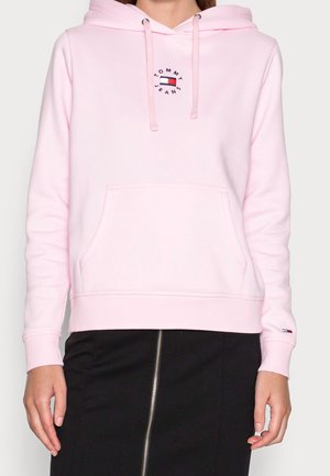 Frau trägt einen hellrosa Hoodie von Tommy Jeans mit Kordelzügen und einer Vorderfronttasche, kombiniert mit einem schwarzen Rock mit einem Reißverschluss vorne.