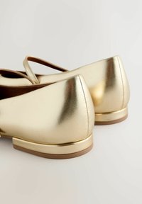 Goldene metallic Ballerinas mit glatter Oberfläche, runder Zehenpartie und niedrigem Absatz. Merkmale ein Überkreuzband-Design nahe der Oberseite. Strukturierte Innenauskleidung.
