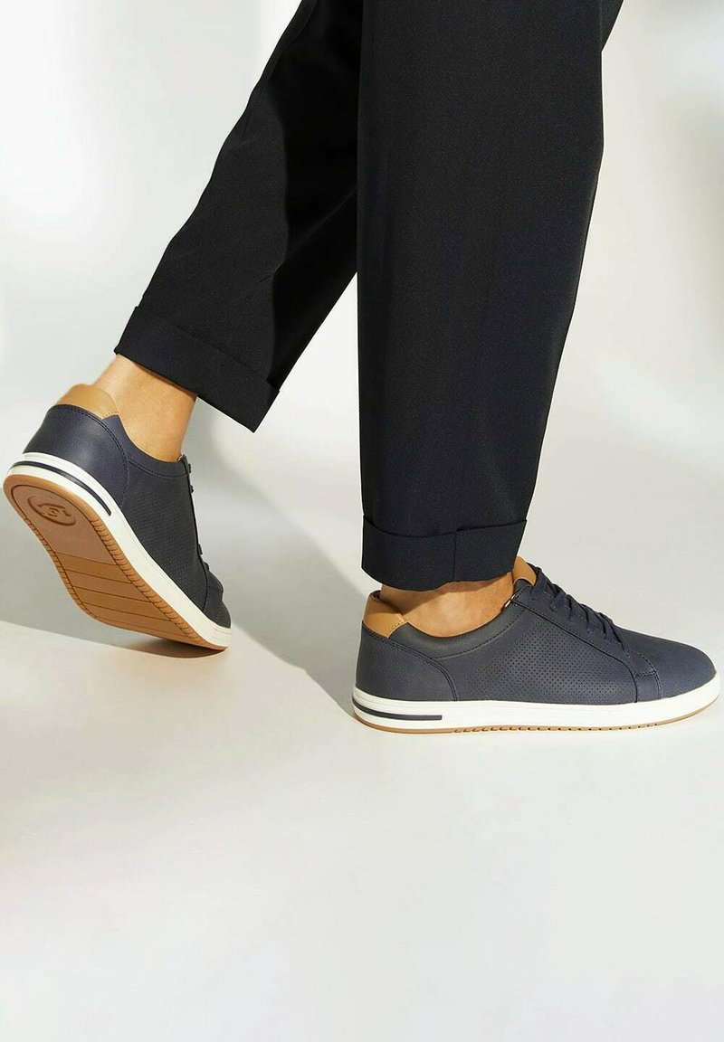 Grey sneaker with perforated design, tan accents, white sole, and rubber tread. Worn with black cuffed trousers, showing side view while walking.