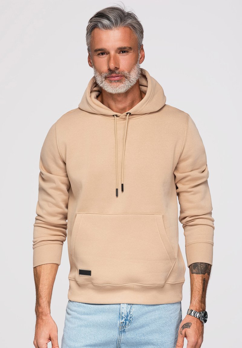 Mann mittleren Alters mit grauen Haaren und Bart, der einen beigen Hoodie und hellblaue Jeans trägt, steht vor einem einfarbigen hellen Hintergrund.