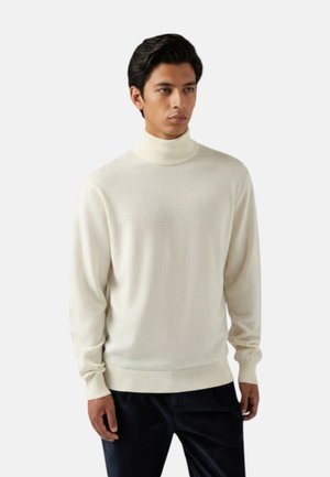 Uomo che indossa un maglione dolcevita semplice color avorio chiaro e pantaloni scuri, in piedi davanti a uno sfondo grigio chiaro con espressione neutra.