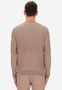 Uomo di spalle che indossa un maglione a coste beige a maniche lunghe e pantaloni beige abbinati su uno sfondo bianco.