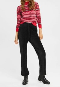 Pull rouge en maille avec motifs géométriques blancs et noirs, associé à un pantalon en velours côtelé noir avec taille à cordon et poches latérales. Bottes noires.