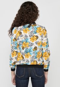 Giacca bomber floreale con base bianca e motivi di ibisco giallo e blu, polsini a costine neri e vestibilità rilassata.