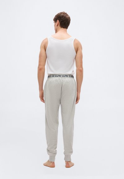 Άνδρας που στέκεται ξυπόλητος, με την πλάτη γυρισμένη, φορώντας λευκό αμάνικο πουκάμισο και ανοιχτό γκρι φόρμα με ζώνη μέσης με λογότυπο.