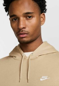 Felpa con cappuccio beige e cordino, realizzata in tessuto morbido. Presenta il logo bianco di Nike sull'area sinistra del petto.