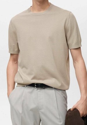 Man met een beige, gebreide top met korte mouwen, ingestopt in lichtgrijze geplooide broek met een zwarte riem, houdt een donkerbruine jas vast.